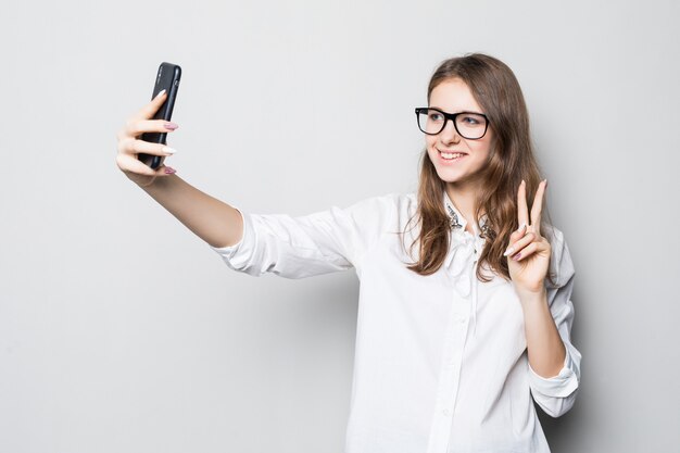 Młoda dziewczyna w okularach ubrana w białą koszulkę ścisłego biura stoi przed białą ścianą i trzyma jej telefon w rękach