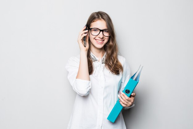 Młoda dziewczyna w okularach ubrana w białą koszulkę ścisłego biura stoi przed białą ścianą i trzyma w rękach jej telefon i teczkę