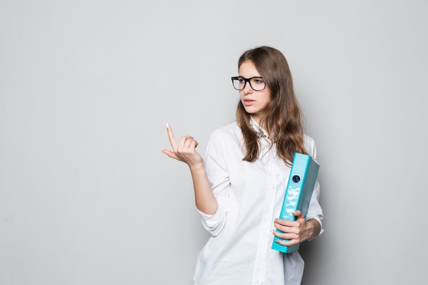 Młoda dziewczyna w okularach ubrana w ścisłej biurowej białej koszulce stoi przed białą ścianą z niebieskim folderem na dokumenty w jej rękach