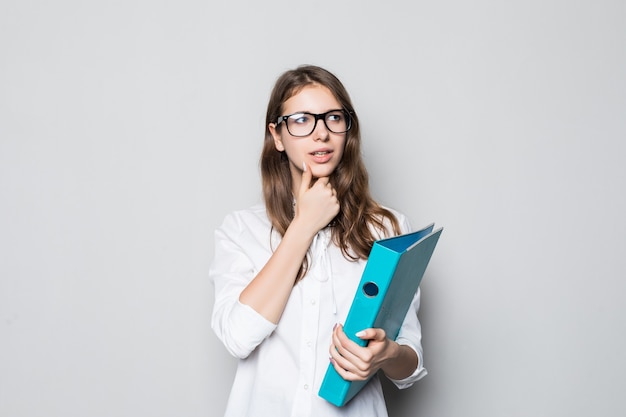 Młoda dziewczyna w okularach ubrana w ścisłej biurowej białej koszulce stoi przed białą ścianą z niebieskim folderem na dokumenty w jej rękach