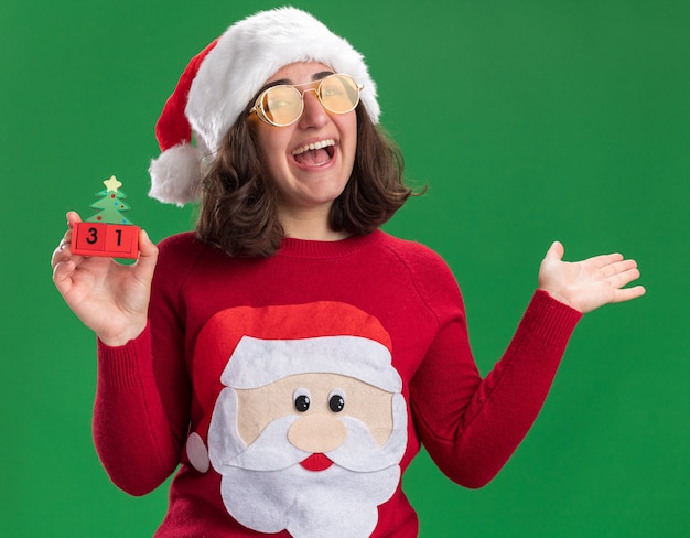 Bezpłatne zdjęcie młoda dziewczyna w świątecznym swetrze w czapce mikołaja i okularach trzymająca kostki z zabawkami z datą nowego roku szczęśliwa i radosna uśmiechnięta podniesiona ręka stojąca nad zieloną ścianą