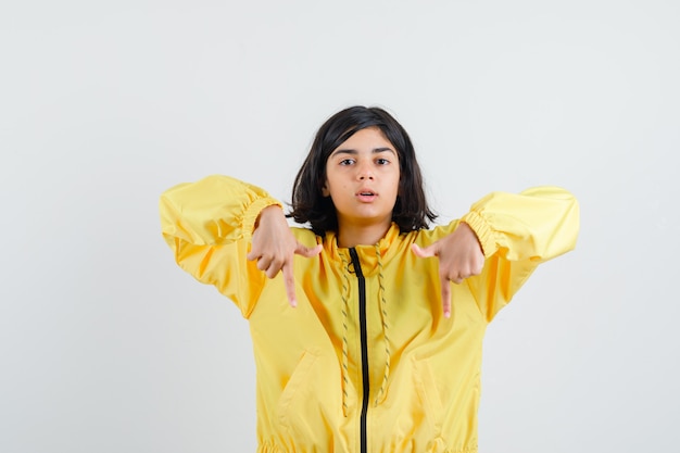 Młoda dziewczyna w żółtej bomber Jacket wskazuje w dół palcami wskazującymi i wygląda poważnie