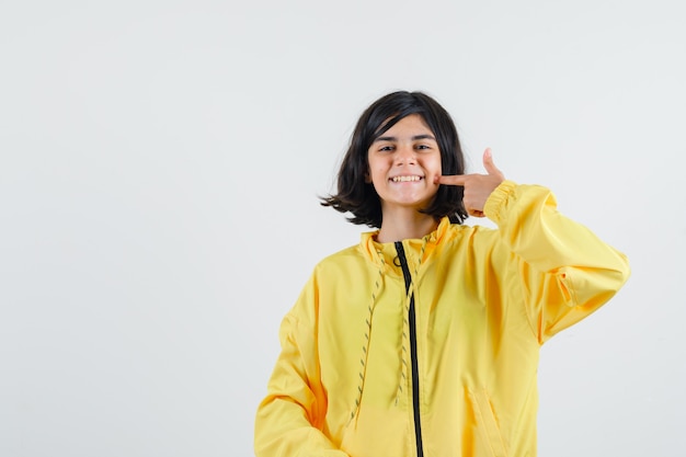 Młoda dziewczyna w żółtej bomber kurtce, uśmiechając się i wskazując palcem wskazującym na policzek i patrząc szczęśliwy
