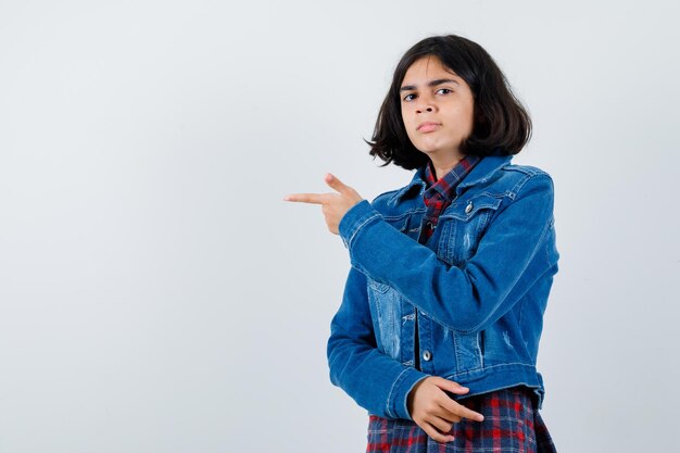 Młoda dziewczyna wskazując w lewo palcem wskazującym w kraciastej koszuli i dżinsowej kurtce i patrząc poważnie, widok z przodu.