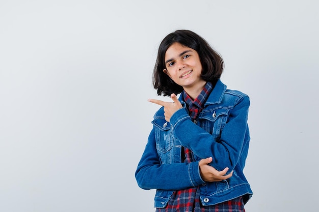Młoda dziewczyna wskazująca w lewo, trzymająca rękę na łokciu w kraciastej koszuli i dżinsowej kurtce i wygląda ładnie.
