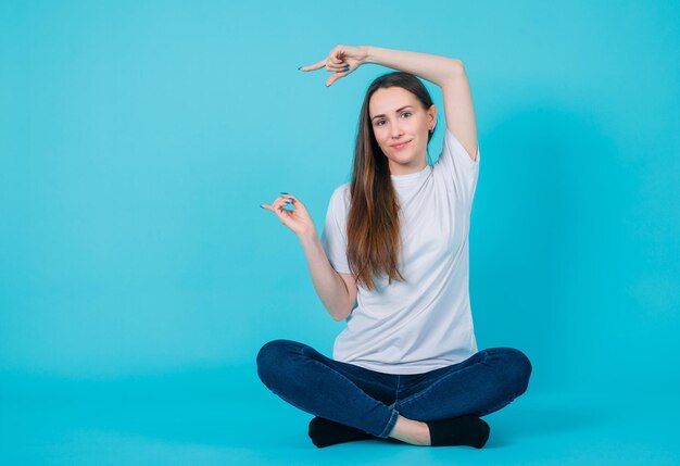 Młoda dziewczyna wskazuje palcami wskazującymi w lewo, siedząc na podłodze na niebieskim tle