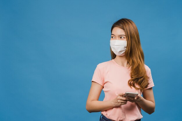 Młoda dziewczyna z Azji nosząca medyczną maskę na twarz, korzystająca z telefonu komórkowego, ubrana w zwykłą odzież odizolowaną na niebieskiej ścianie
