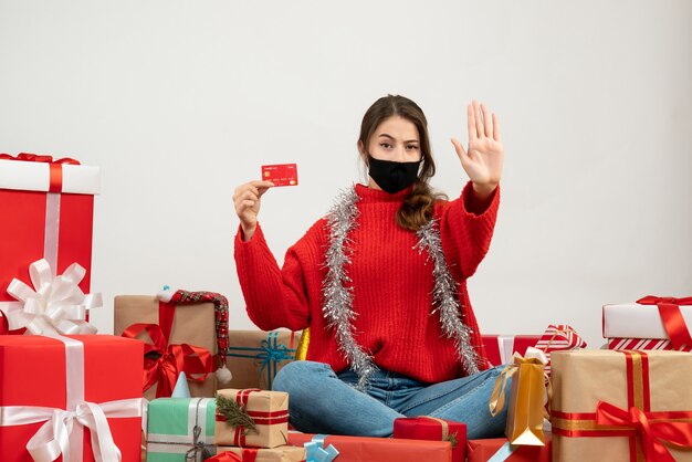 młoda dziewczyna z czerwonym swetrem i czarną maską trzymając kartę siedzi wokół prezentów na białym tle