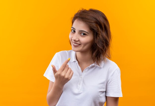 Młoda dziewczyna z krótkimi włosami na sobie białą koszulkę polo, uśmiechając się z radosna buźka pokazuje palec wskazujący
