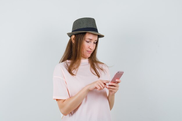 Młoda dziewczyna za pomocą telefonu komórkowego w różowy t-shirt, kapelusz i patrząc zajęty, widok z przodu.
