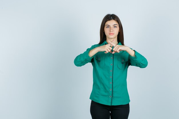 Bezpłatne zdjęcie młoda dziewczyna zderzająca się z palcami wskazującymi, gestykulująca x podpisuje się w zielonej bluzce, czarnych spodniach i wygląda na szczęśliwą. przedni widok.