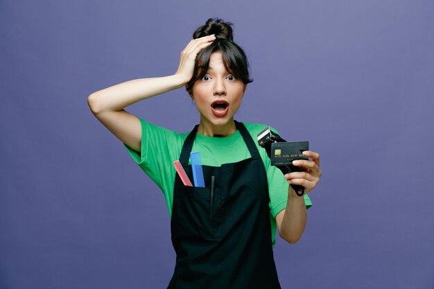 Młoda fryzjerka kobieta nosi fartuch, trzymając golarkę elektryczną i kartę kredytową, patrząc na aparat zmartwiony i zdezorientowany, trzymając rękę na głowie za błąd stojący na niebieskim tle