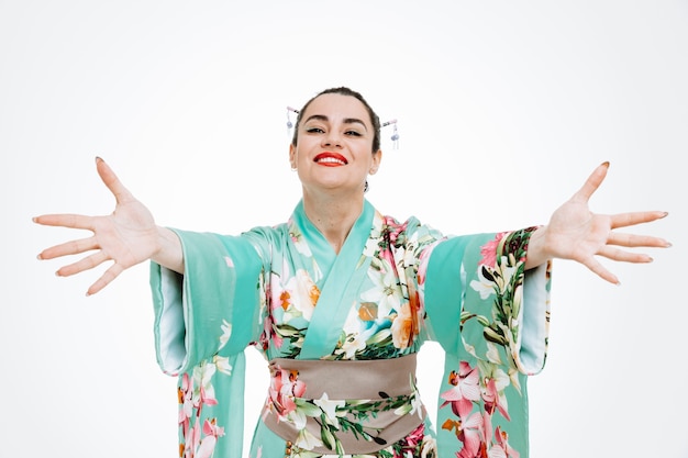 młoda gejsza kobieta w tradycyjnym japońskim kimonie patrząca z przodu uśmiechnięta radośnie wykonująca powitalny gest szeroko otwierające się ręce stojące nad białą ścianą