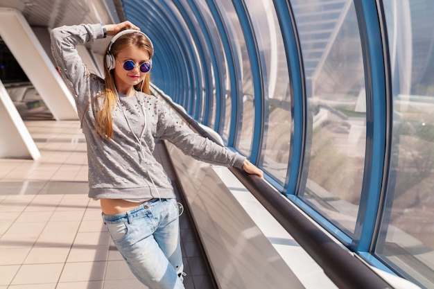 Bezpłatne zdjęcie młoda hipster kobieta tańczy, zabawy w miejskim nowoczesnym budynku ubrana w strój codzienny, słuchając muzyki w słuchawkach, ubrana w dżinsy, sweter i okulary przeciwsłoneczne