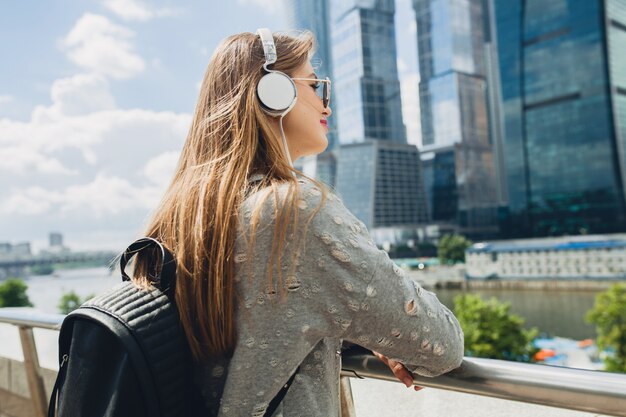Młoda hipster kobieta, zabawy na ulicy, słuchanie muzyki na słuchawkach, na sobie różowe okulary i plecak, miejski styl wiosna lato