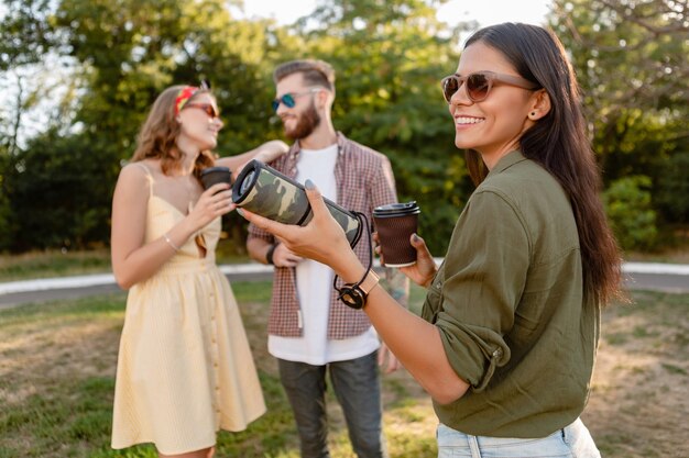 Młoda hipsterska firma przyjaciół, którzy wspólnie bawią się w parku, uśmiechając się, słuchając muzyki na bezprzewodowym głośniku w sezonie letnim