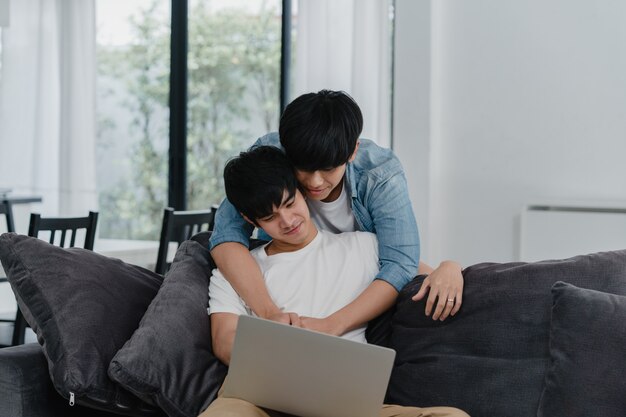 Młoda Homoseksualna para używa komputerowego laptop w nowożytnym domu. Azjaci LGBTQ + mężczyźni chętnie relaksują się przy użyciu technologii, oglądając wspólnie film w Internecie, leżąc na kanapie w salonie w domu.