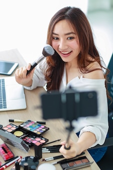 Młoda i urocza azjatycka vlogerka, influencerka lub sprzedawczyni online trzyma smartfon na długim kiju, aby zrobić sobie wideo selfie i transmitować na żywo do recenzji kosmetyków. koncepcja marketingu online.
