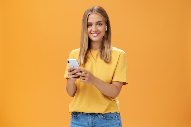 Młoda kaukaska dziewczyna o opalonej skórze i jasnych włosach, korzystająca z bezprzewodowych słuchawek, aby zadzwonić do przyjaciela przez smartfona, trzymając telefon przy klatce piersiowej, uśmiechając się radośnie do kamery, przyzwyczajając się do nowej technologii