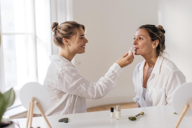 Młoda Kaukaska Dziewczyna Powącha Serum Do Twarzy Dorosłej Kobiecie Siedzącej Przed Nią Przy Stole W Pomieszczeniu. Koncepcja Domowego Spa Na Twarz