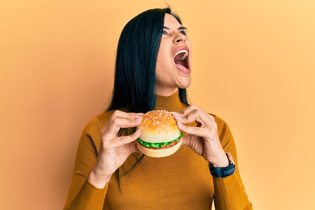 Bezpłatne zdjęcie młoda kaukaska kobieta jedząca smacznego klasycznego burgera wściekła i szalona krzycząca sfrustrowana i wściekła krzycząca ze złością patrzącą w górę