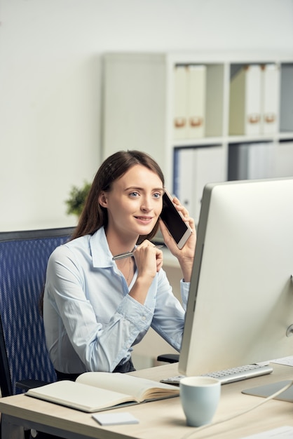 Młoda Kaukaska kobieta siedzi w biurze przed ekranem komputerowym i opowiada na telefonie