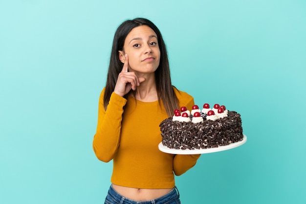 Młoda kaukaska kobieta trzyma tort urodzinowy na białym tle na niebieskim tle, myśląc o pomyśle