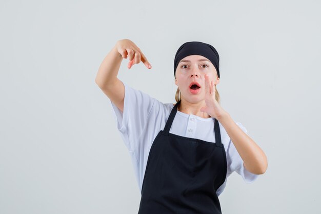 Młoda kelnerka wskazująca na podniesioną dłoń w mundurze i fartuchu i wyglądająca na zaskoczoną