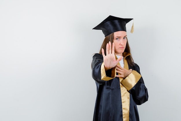 Bezpłatne zdjęcie młoda kobieta absolwentka pokazując gest stop w akademickim stroju i patrząc poważny, widok z przodu.