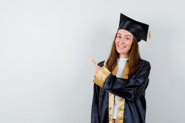 Młoda kobieta absolwentka w akademickim stroju, wskazując na lewy górny róg i patrząc szczęśliwy, widok z przodu.