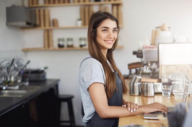 Młoda kobieta barista w swoim miejscu pracy wygląda na szczęśliwą, uśmiechniętą, pracującą na laptopie
