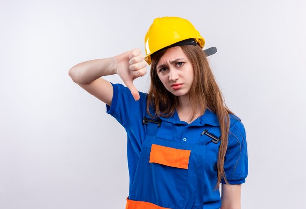 Młoda kobieta budowniczy pracownik w mundurze budowlanym i hełmie ochronnym, patrząc niezadowolony, pokazując kciuk w dół stojąc na białej ścianie
