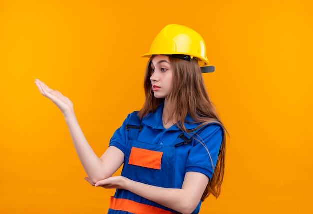 Młoda kobieta budowniczy pracownik w mundurze budowy i hełmie ochronnym patrząc na bok niezadowolony podnosząc rękę, argumentując stojąc nad pomarańczową ścianą