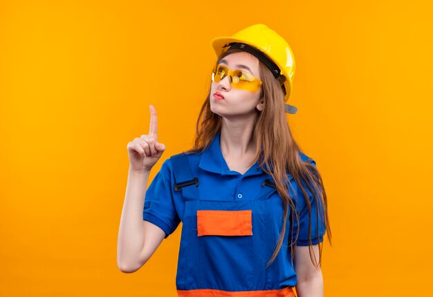 Młoda kobieta budowniczy w mundurze budowlanym i kasku, patrząc zaintrygowany, wskazując palcem wskazującym na coś stojącego