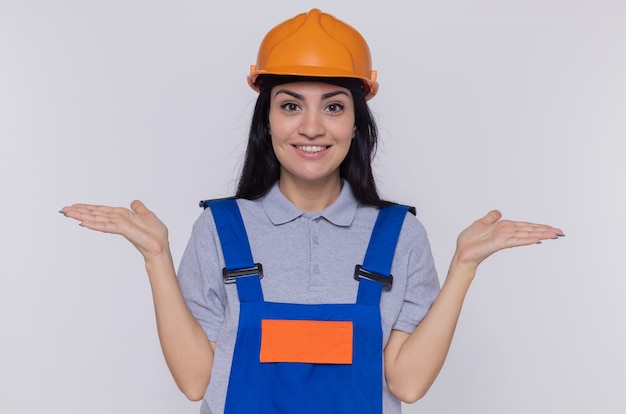 Młoda kobieta budowniczy w mundurze konstrukcyjnym i kasku ochronnym patrząc na przód uśmiechnięty, rozkładając ramiona na boki stojąc nad białą ścianą
