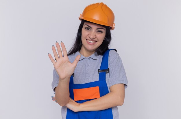 Młoda kobieta budowniczy w mundurze konstrukcyjnym i kasku ochronnym patrząc z przodu ze sceptycznym uśmiechem na twarzy pokazując otwartą dłoń stojącą nad białą ścianą