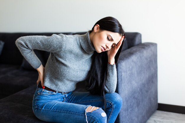 Młoda Kobieta cierpi na ból pleców i narzeka, siedząc na kanapie w salonie w domu