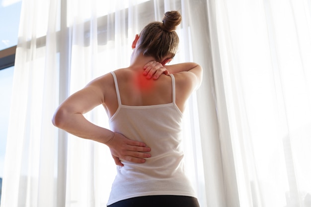 Młoda kobieta cierpi na ból szyi i bóle pleców, rozciągając mięśnie w domu. ból pleców i szyi kobiety