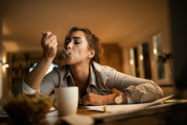 Młoda kobieta czuje głód i je sałatkę podczas wieczornej nauki w domu