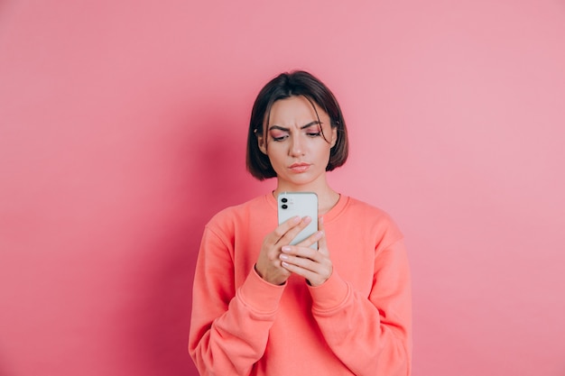 Młoda kobieta czuje się smutny niezadowolony zły podczas czytania sms przy użyciu swojego telefonu komórkowego, na różowym tle