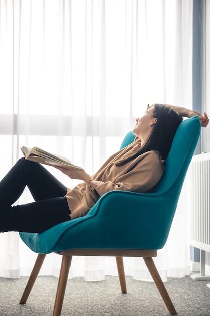 Bezpłatne zdjęcie młoda kobieta czyta książkę siedząc na fotelu przy oknie