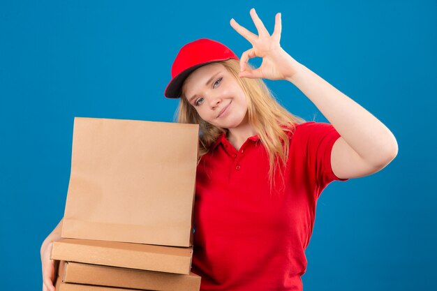 Młoda kobieta dostawy na sobie czerwoną koszulkę polo i czapkę stojącą z pakietem papieru robi ok znak uśmiechnięty przyjazny na odosobnionym niebieskim tle
