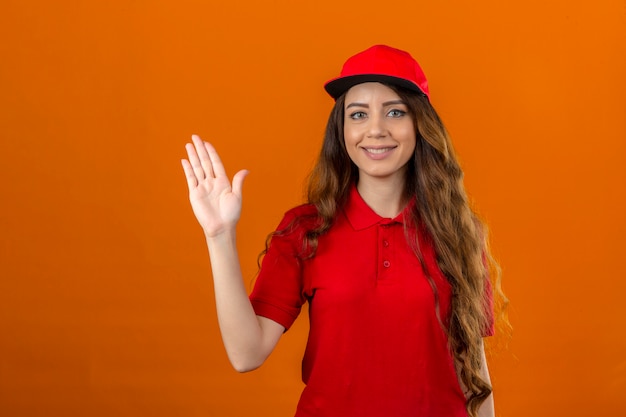Młoda kobieta dostawy ubrana w czerwoną koszulkę polo i czapkę, uśmiechnięta radośnie i wesoło machająca ręką, witająca i witająca lub żegnająca się na odizolowanym pomarańczowym tle