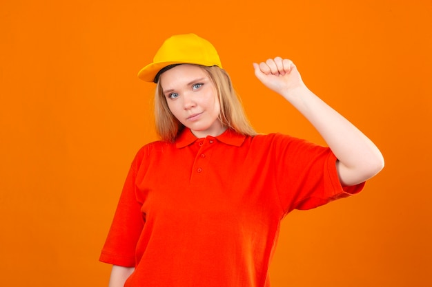 Młoda kobieta dostawy ubrana w czerwoną koszulkę polo i żółtą czapkę uśmiechnięta przyjazna pozycja z podniesioną pięścią zwycięzcę koncepcji na białym tle pomarańczowy