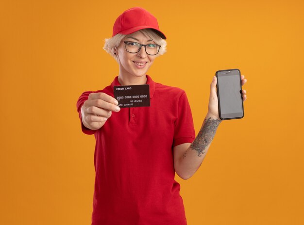 Młoda kobieta dostawy w czerwonym mundurze i czapce w okularach, pokazując smartfon i kartę kredytową, uśmiechając się pewnie stojąc na pomarańczowej ścianie