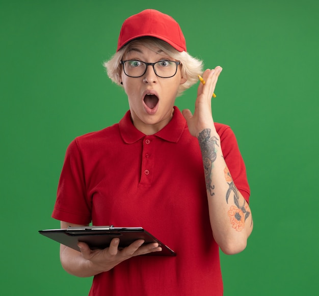 Młoda kobieta dostawy w czerwonym mundurze i czapce w okularach ze schowkiem z pustymi stronami i ołówkiem wyglądająca na zdumioną i zaskoczoną stojącą nad zieloną ścianą