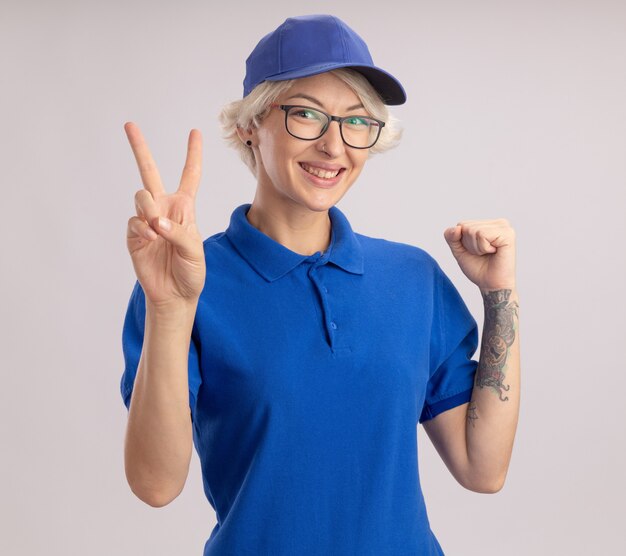 Młoda kobieta dostawy w niebieskim mundurze i czapce patrząc podnosząc pięść pokazując znak v uśmiechnięty wesoło stojąc na białej ścianie