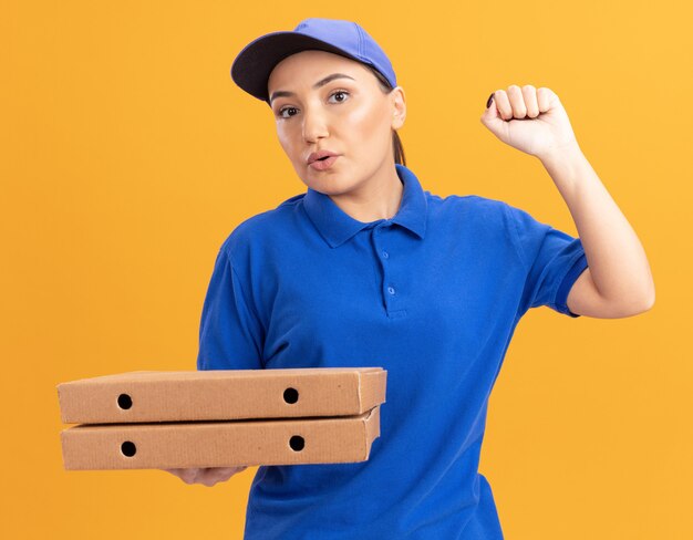 Młoda kobieta dostawy w niebieskim mundurze i czapce trzyma pudełka po pizzy patrząc z przodu z pewnym siebie wyrazem podnosząc pięść stojącą nad pomarańczową ścianą