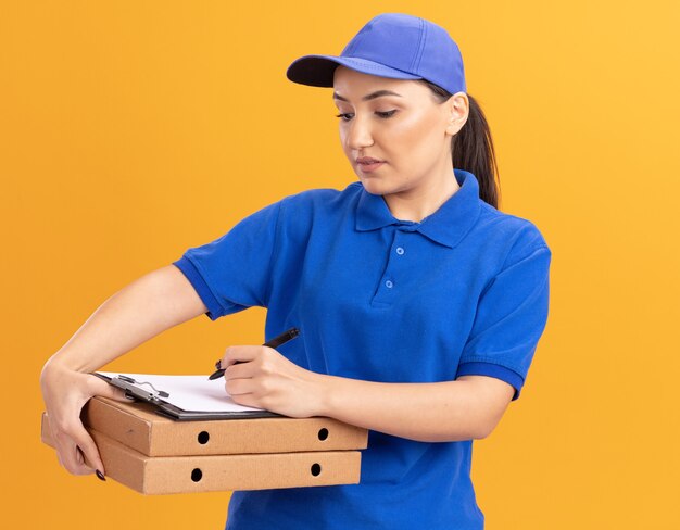 Młoda kobieta dostawy w niebieskim mundurze i czapce, trzymając pudełka po pizzy i pisanie w schowku z poważną twarzą stojącą nad pomarańczową ścianą