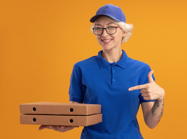 Młoda kobieta dostawy w niebieskim mundurze i czapce w okularach, trzymając pudełka po pizzy, wskazując palcem wskazującym na nich, uśmiechając się wesoło nad pomarańczową ścianą